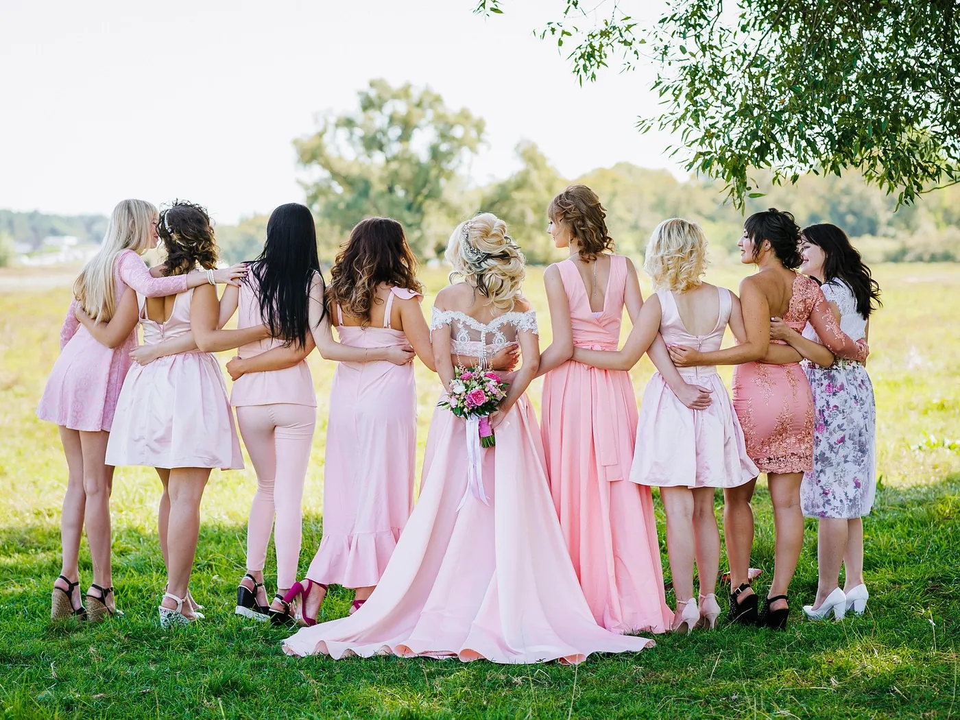 Bride and bridesmaids in pink dresses standing arm in arm in a meadow
